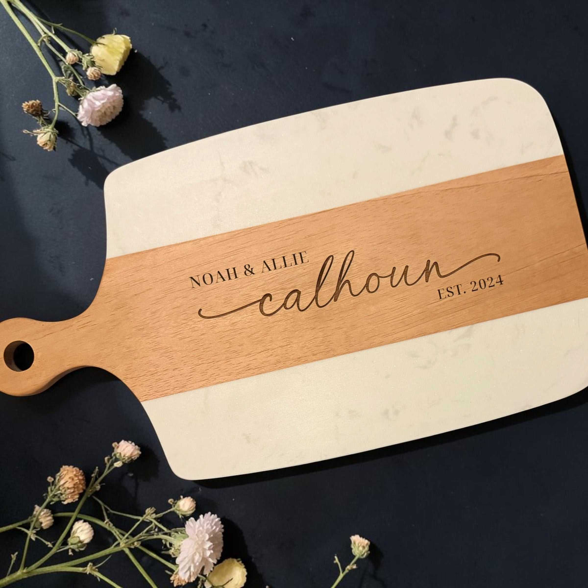 The Storyboard wood and white marble serving board with a personalized engraved name, date, and hanging handle